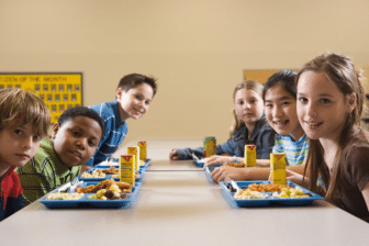 A group of five school-aged children eating lunch at a cafeteria table and smiling to the camera.