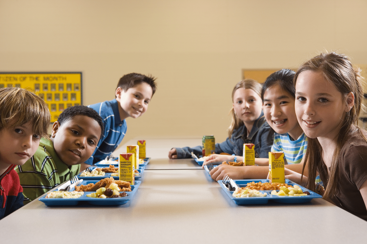 A group of five school-aged children eating lunch at a cafeteria table and smiling to the camera.